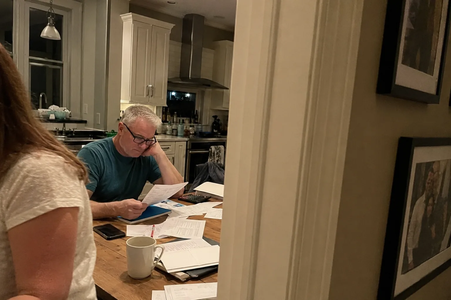 Illustrative editorial photo: evening kitchen table with bills, papers, coffee and a phone in a tired domestic setting.